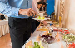Mężczyna nakładający sobie przekąski na talerz ze stołu cateringowego zastawionego półmiskami.