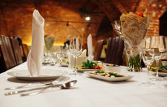 Piwnica restauracyjna z ceglanymi ścianami i nastrojowym oświetleniem. Przybrany na biało stół z zastawą i talerzami z przekąskami.