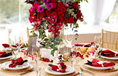 Okrągły stolik na 8 osób z białym serwisem i ciemno-czerwonymi serwetkami na talerzach. Na środku bukiet czerwonych kwiatów.