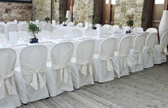 Duży stół udekorowany na biało w sali ślubnej. Na krzesłach białe draperie.