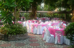 Zielony zacieniony ogród z okrągłymi stołami z różowymi obrusami i białą zastawą. Na krzesłach biały materiał przewiazany różowymi wstążkami.