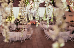 Sala z okrągłymi stołami z biała zastawą. Z sufitu zwisają girlandy białych kwiatów.